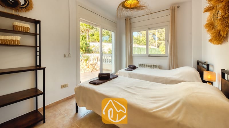 Dormitorio con dos camas individuales, ropa de cama en tonos neutros, estantería de madera oscura y puertas corredizas de cristal que se abren a una soleada terraza.
