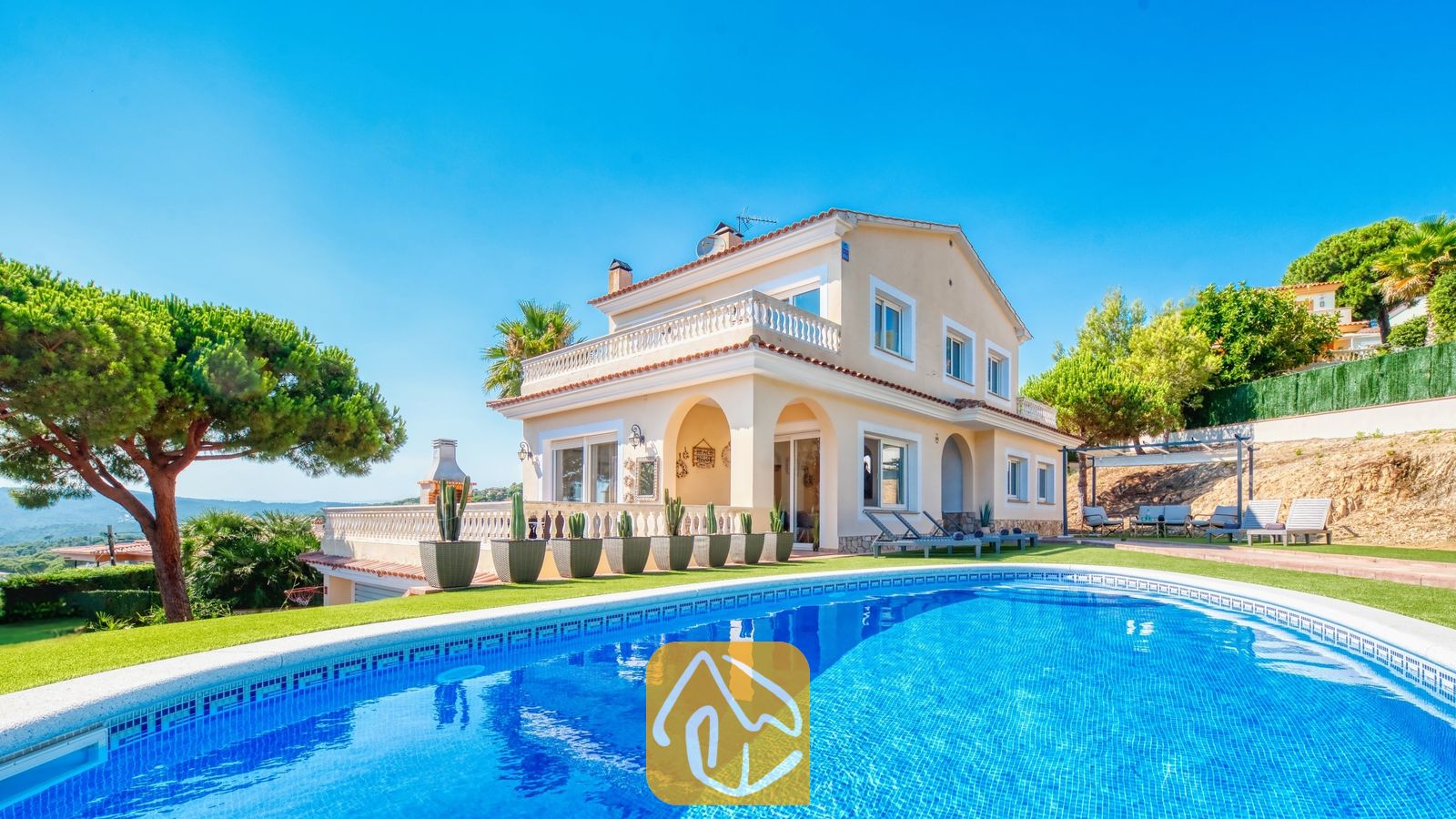 Piscina privada en primer plano con la Villa Ibiza, una casa de dos plantas de color crema, césped verde y cactus en macetas bajo un cielo azul despejado.