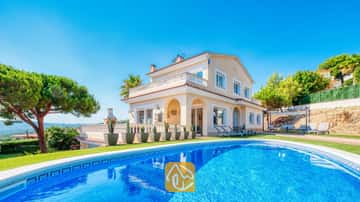 Piscina privada en primer plano con la Villa Ibiza, una casa de dos plantas de color crema, césped verde y cactus en macetas bajo un cielo azul despejado.