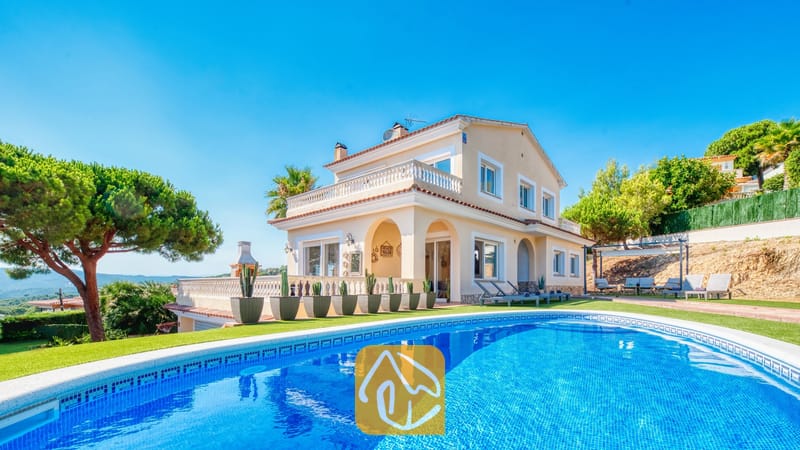 Piscina privada en primer plano con la Villa Ibiza, una casa de dos plantas de color crema, césped verde y cactus en macetas bajo un cielo azul despejado.