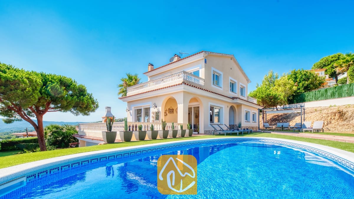 Piscina privada en primer plano con la Villa Ibiza, una casa de dos plantas de color crema, césped verde y cactus en macetas bajo un cielo azul despejado.