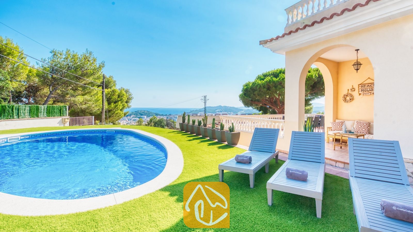 Piscina privada y tumbonas de madera azul sobre césped artificial en Villa Ibiza, con vistas al mar lejano y a los árboles.