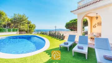 Piscina privada y tumbonas de madera azul sobre césped artificial en Villa Ibiza, con vistas al mar lejano y a los árboles.