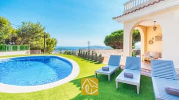 Piscina privada y tumbonas de madera azul sobre césped artificial en Villa Ibiza, con vistas al mar lejano y a los árboles.