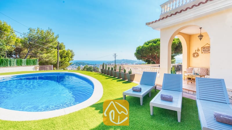 Piscina privada y tumbonas de madera azul sobre césped artificial en Villa Ibiza, con vistas al mar lejano y a los árboles.