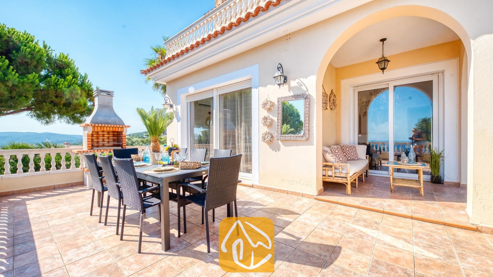 Terraza exterior en la Villa Ibiza con una mesa de comedor, sillas negras, una barbacoa de obra y una zona de estar cubierta bajo un porche de medio punto.