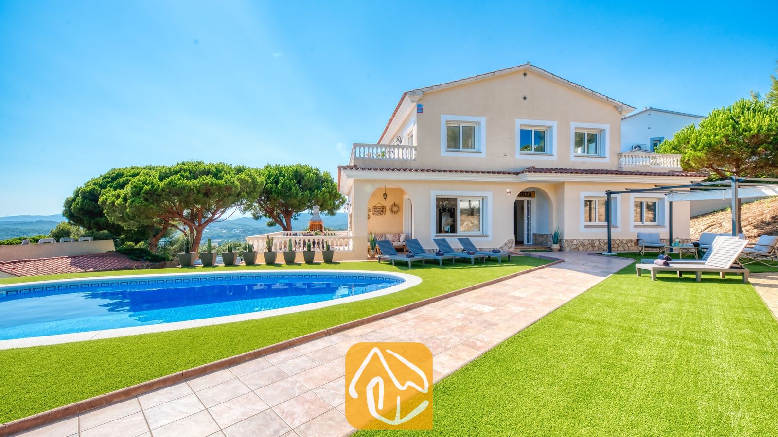 Vista exterior de Villa Ibiza que muestra una piscina azul de forma curva, césped artificial verde, tumbonas y una casa beige de dos plantas bajo un cielo azul despejado.