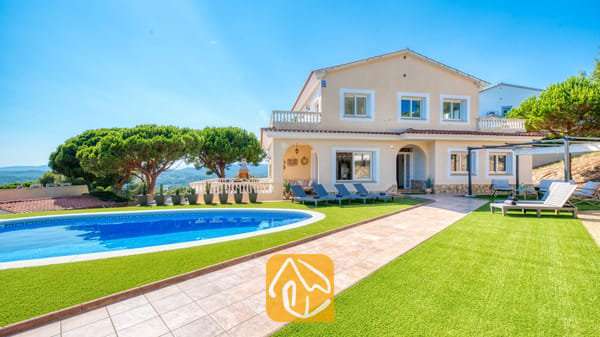 Vista exterior de Villa Ibiza que muestra una piscina azul de forma curva, césped artificial verde, tumbonas y una casa beige de dos plantas bajo un cielo azul despejado.