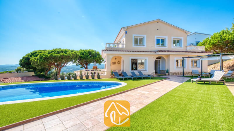 Vista exterior de Villa Ibiza que muestra una piscina azul de forma curva, césped artificial verde, tumbonas y una casa beige de dos plantas bajo un cielo azul despejado.