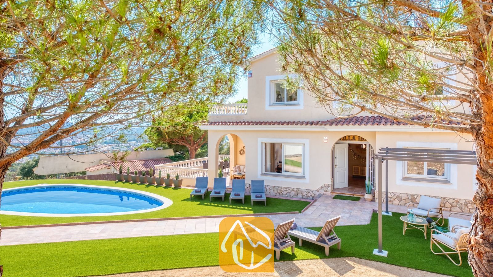 Vista exterior de Villa Ibiza que muestra una piscina de diseño curvo, un césped artificial, múltiples tumbonas grises y una casa beige de dos plantas con tejas de terracota.