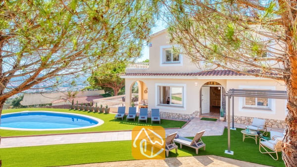 Vista exterior de Villa Ibiza que muestra una piscina de diseño curvo, un césped artificial, múltiples tumbonas grises y una casa beige de dos plantas con tejas de terracota.