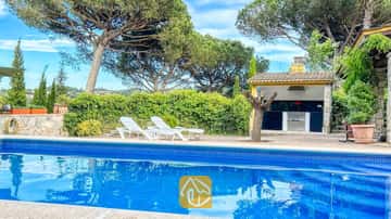 Piscina privada con borde de azulejos azules y tumbonas blancas en la terraza de Villa Iako, que cuenta con una zona de barbacoa al aire libre y altos pinos al fondo.