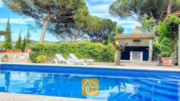 Piscina privada con borde de azulejos azules y tumbonas blancas en la terraza de Villa Iako, que cuenta con una zona de barbacoa al aire libre y altos pinos al fondo.