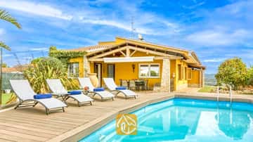 Piscina privada con tarima de madera y tumbonas blancas en Villa Soleia, España, que cuenta con una villa de exterior amarillo y cielo azul.