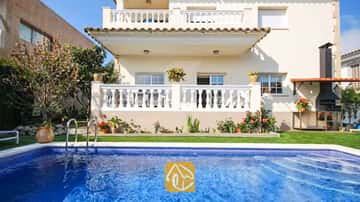 Piscina privada de aguas azuladas frente a la fachada exterior de Villa Mercedes Fenals, que cuenta con balcones de balaustrada blanca, césped y barbacoa de obra en el exterior.