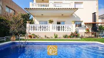 Piscina privada de aguas azuladas frente a la fachada exterior de Villa Mercedes Fenals, que cuenta con balcones de balaustrada blanca, césped y barbacoa de obra en el exterior.