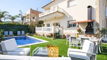 Vista exterior de la Villa Mercedes Fenals que muestra una piscina, césped, mobiliario de exterior blanco y una barbacoa de obra integrada contra la fachada de tonos crema.